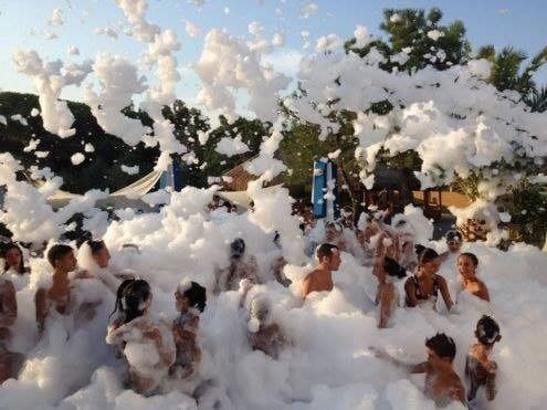 Folla a una festa all'aperto piena di schiuma, con persone che ridono sotto gli spruzzi di schiuma bianca.