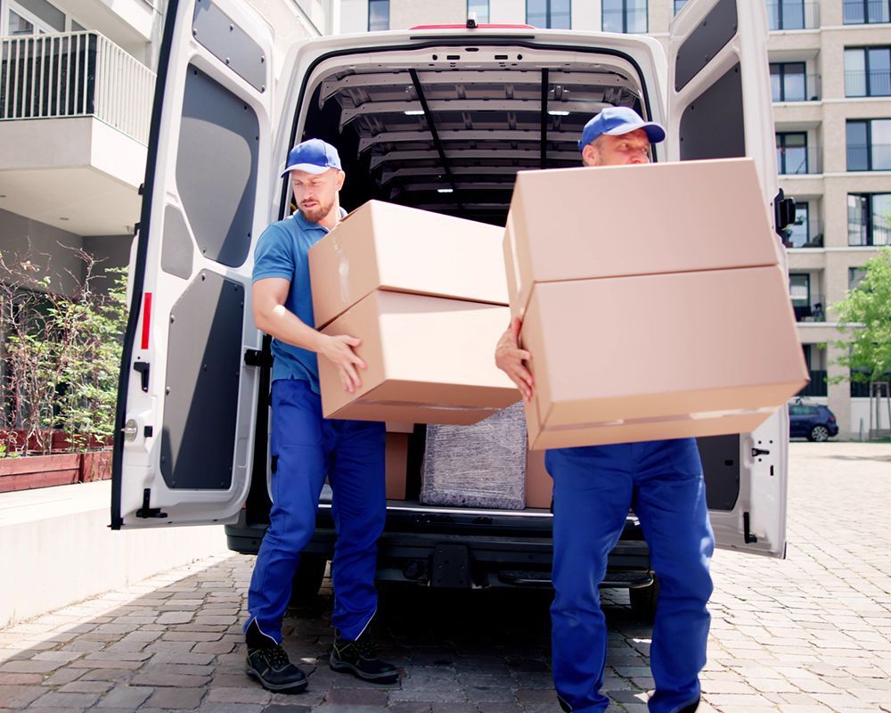 Two movers in blue uniforms unload boxes from a white van on a sunny day.