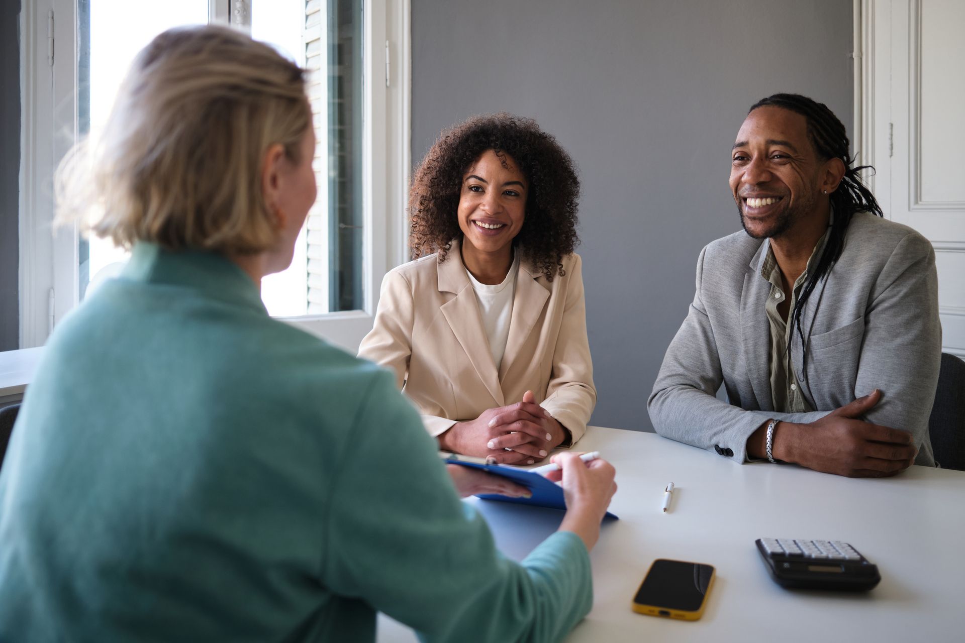 Woman in turquoise jacket speaks to a smiling couple at a table in an office.