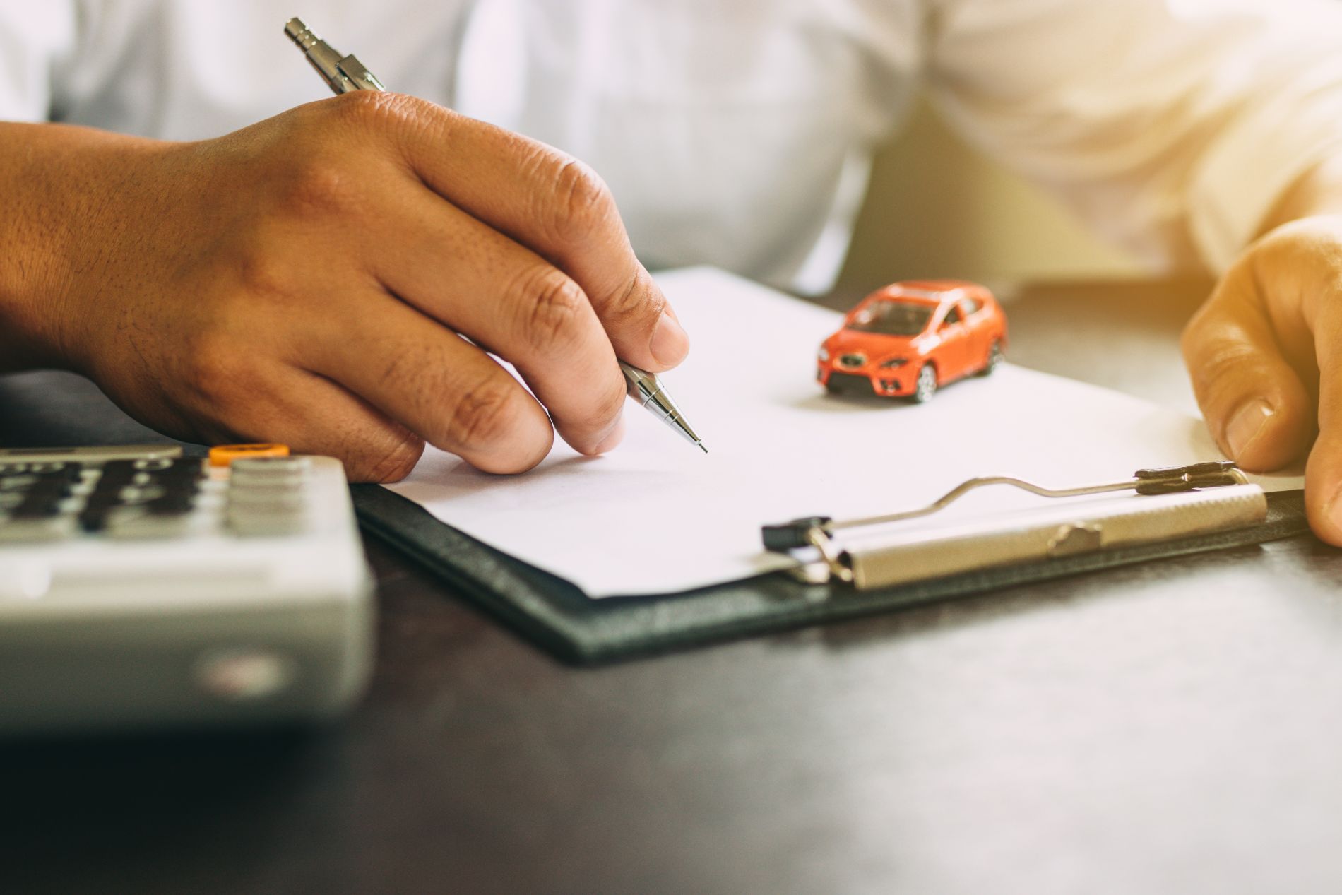 Person writing on paper, next to a toy car and calculator, possibly completing paperwork.