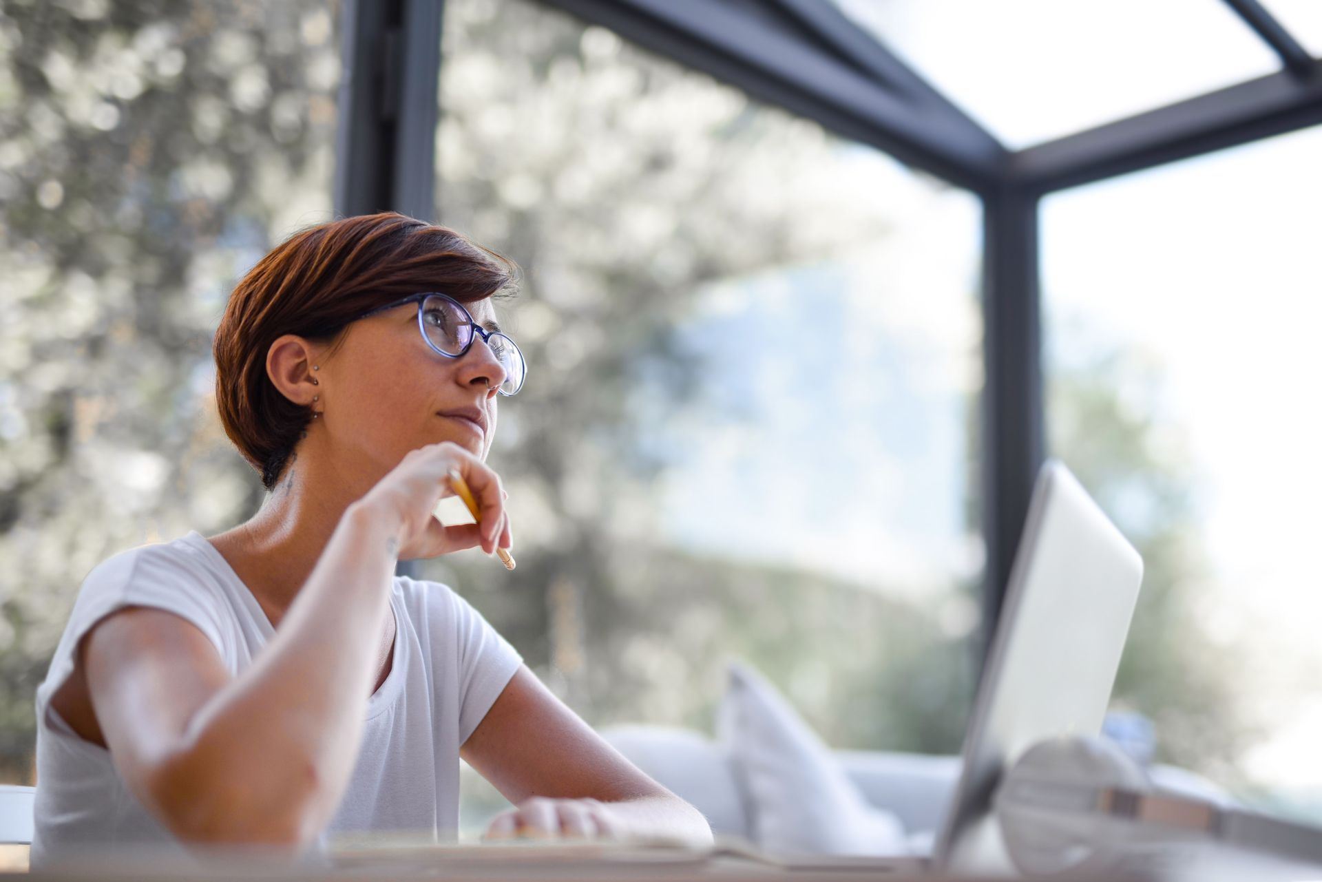 Person in glasses drinks from a cup while working on a laptop outdoors under a shaded patio