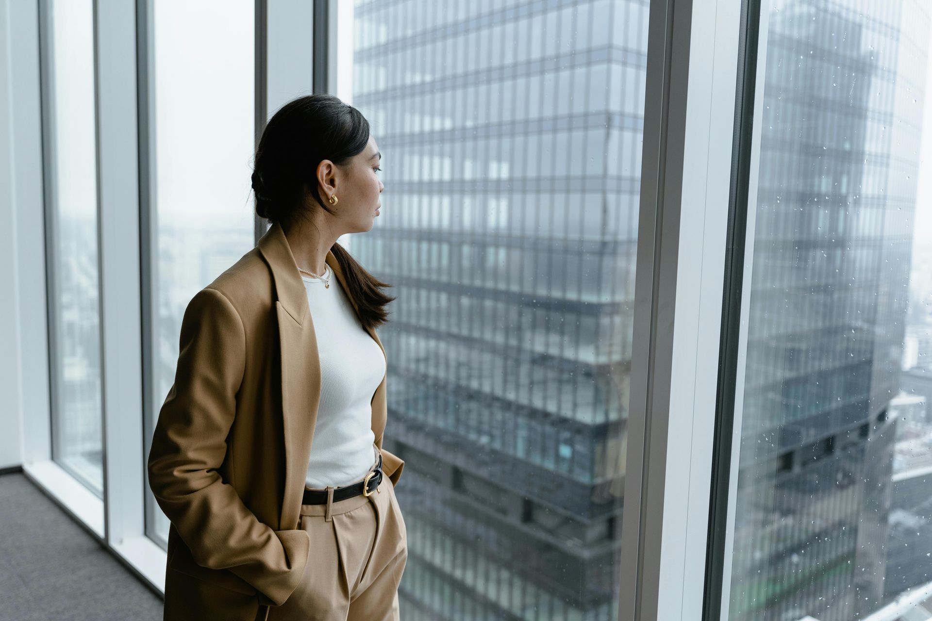 Woman in a tan suit looking out a window at tall office buildings