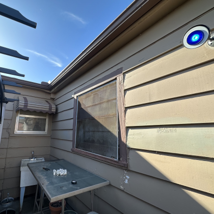 A brown house exterior with a window, a sink, a table with small white objects, and a camera with a glowing blue light.