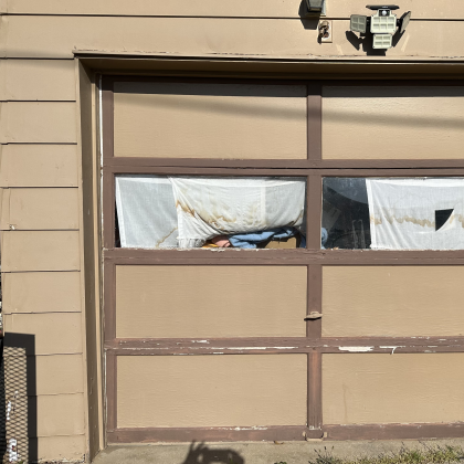 A person rests inside a garage, visible through the paneled door's window, which is partially covered by a white sheet.