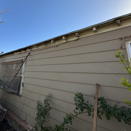 An exterior wall of a house with missing fascia boards, revealing exposed rafters under a clear blue sky.