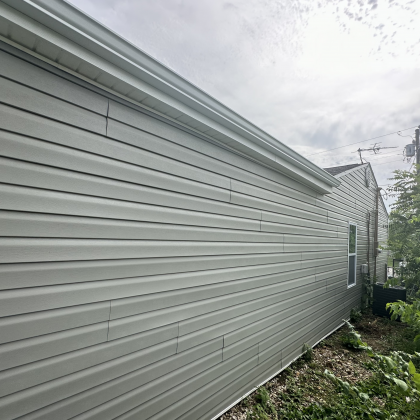 The side of a house with light green vinyl siding and an overcast sky, showing a seam line running down the wall.