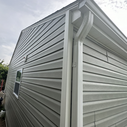 Light gray vinyl siding on the corner of a building with a white metal gutter and downspout under a cloudy sky.