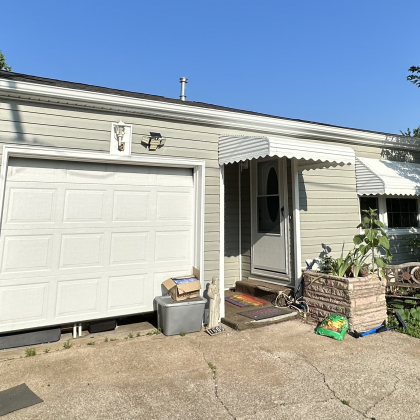 A light-colored house with a single-car garage, a white front door under a small awning, and a planter with greenery.