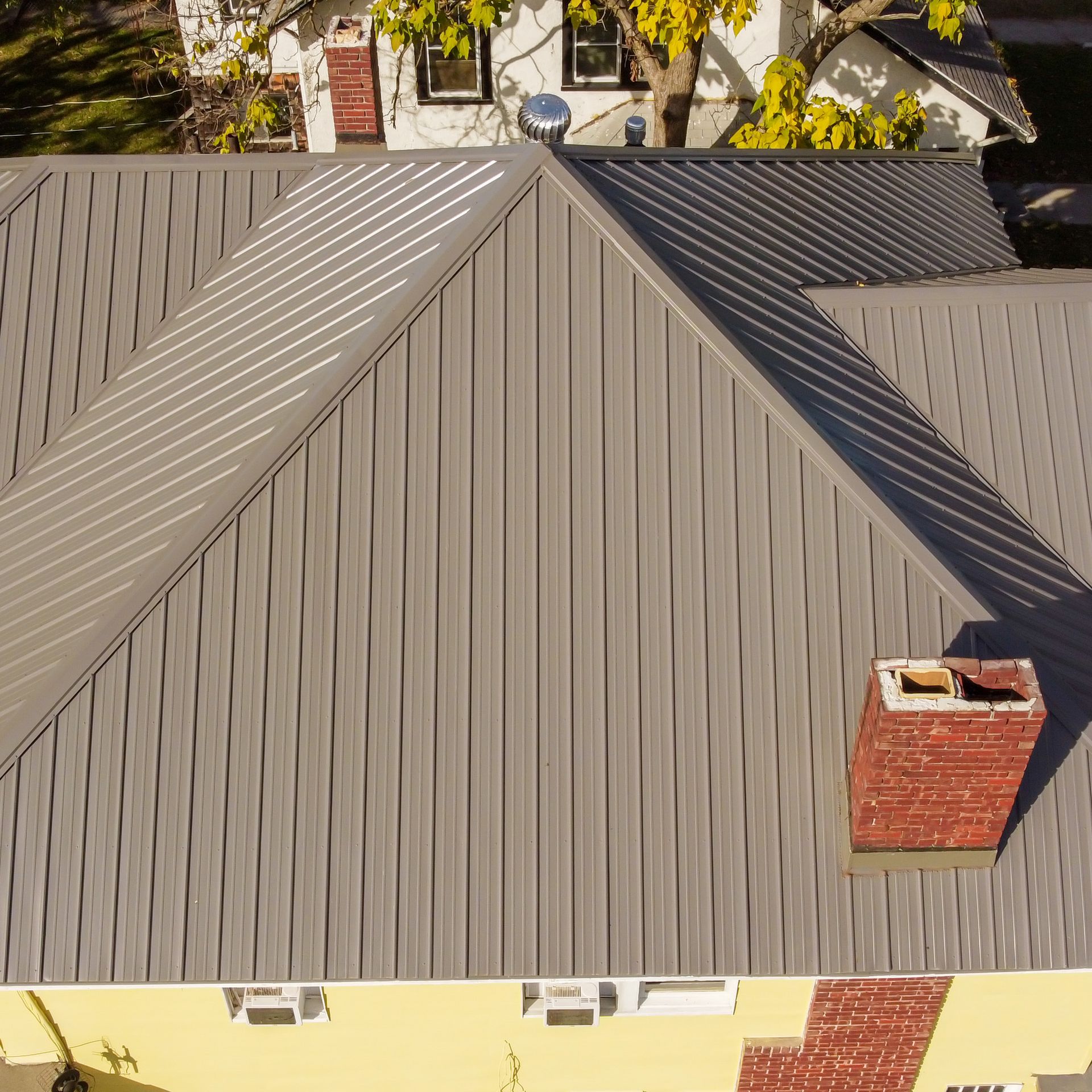 An aerial view of a gray metal roof with standing seams, a brick chimney, and a small silver vent.