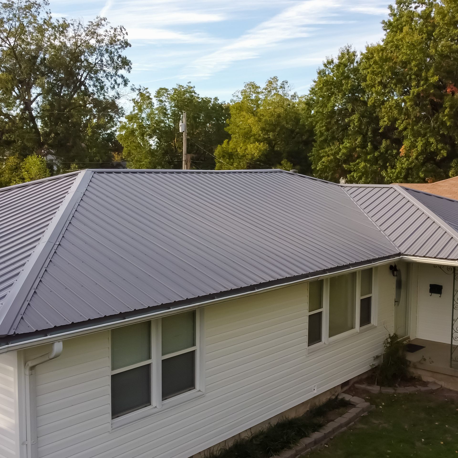 A side view of a white, single-story house with a new grey metal roof and white siding, set against green trees.