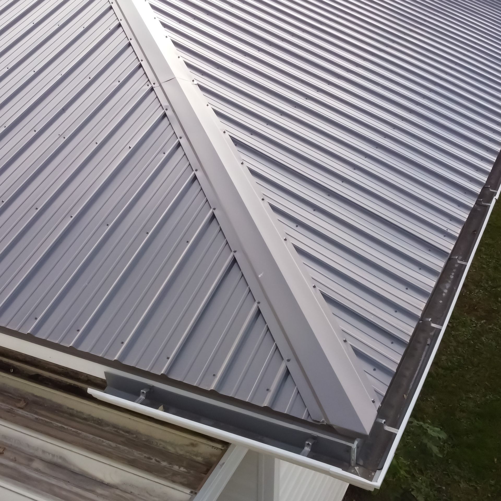 A close-up view of a metal roof with a white ridge cap, dark metal trim, and a gutter system on the edge.
