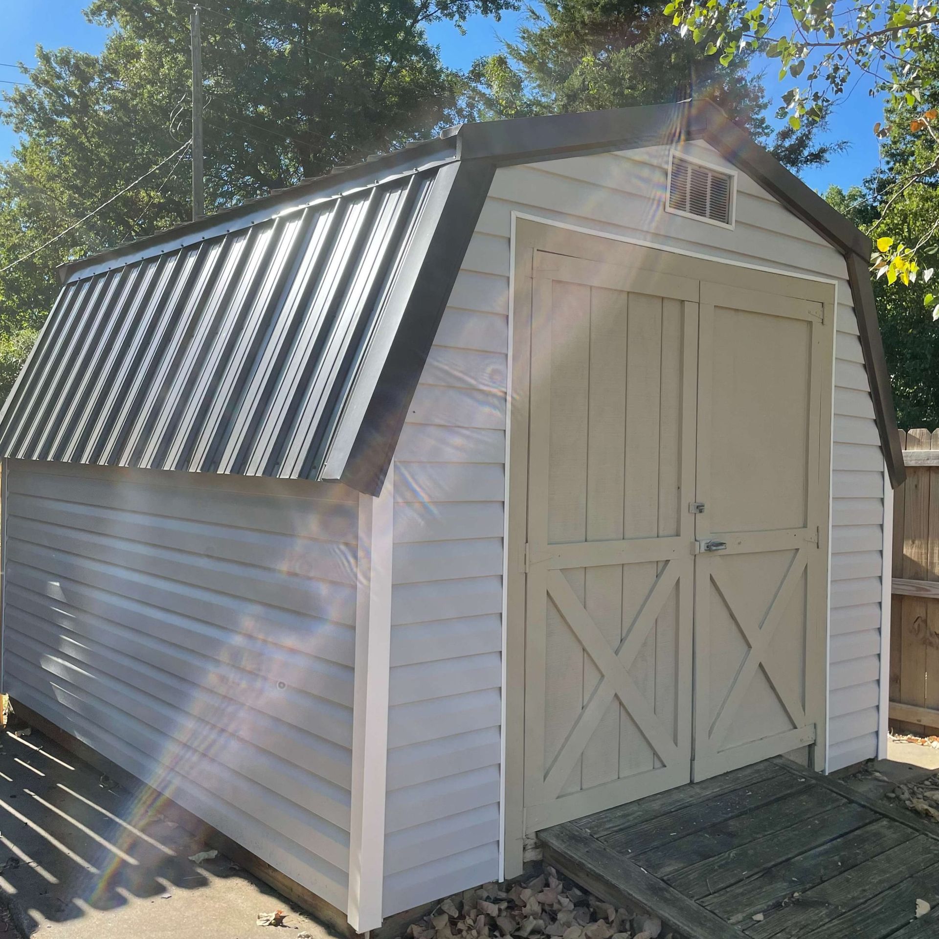 A light-gray shed with double doors and a dark metal gambrel roof stands outdoors on a sunny day.