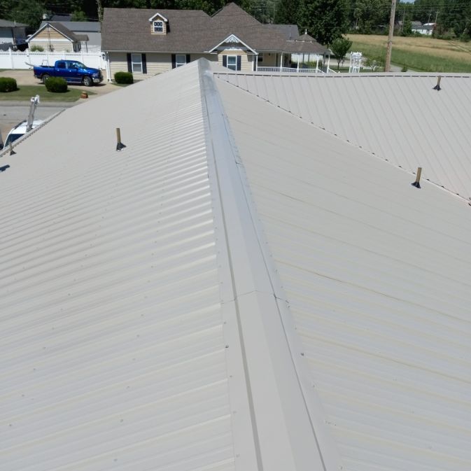 A high-angle view of a beige metal roof featuring a central ridge cap, with a house and blue truck visible below.