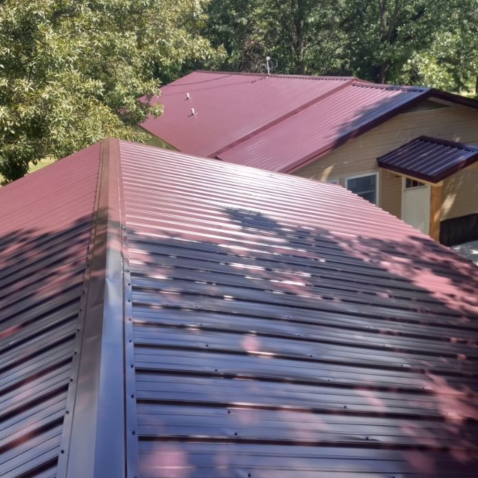 A high-angle view of a house with a dark red metal roof, partially shaded by trees.