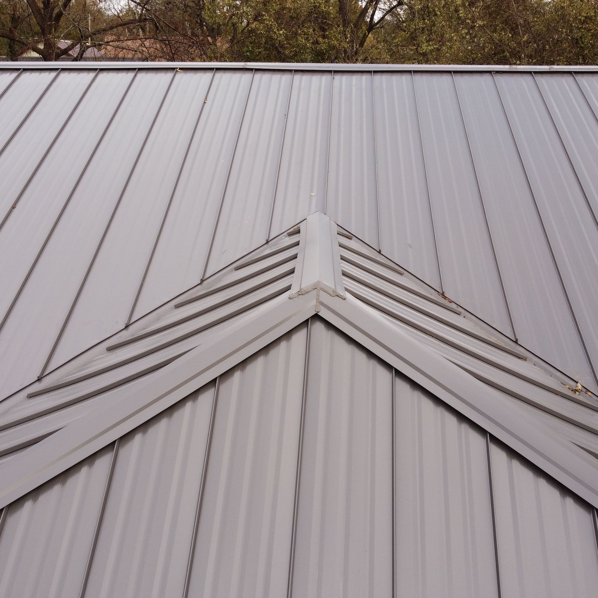 A close-up view of a metal roof with light gray standing seam panels, featuring a triangular ridge cap and peak.