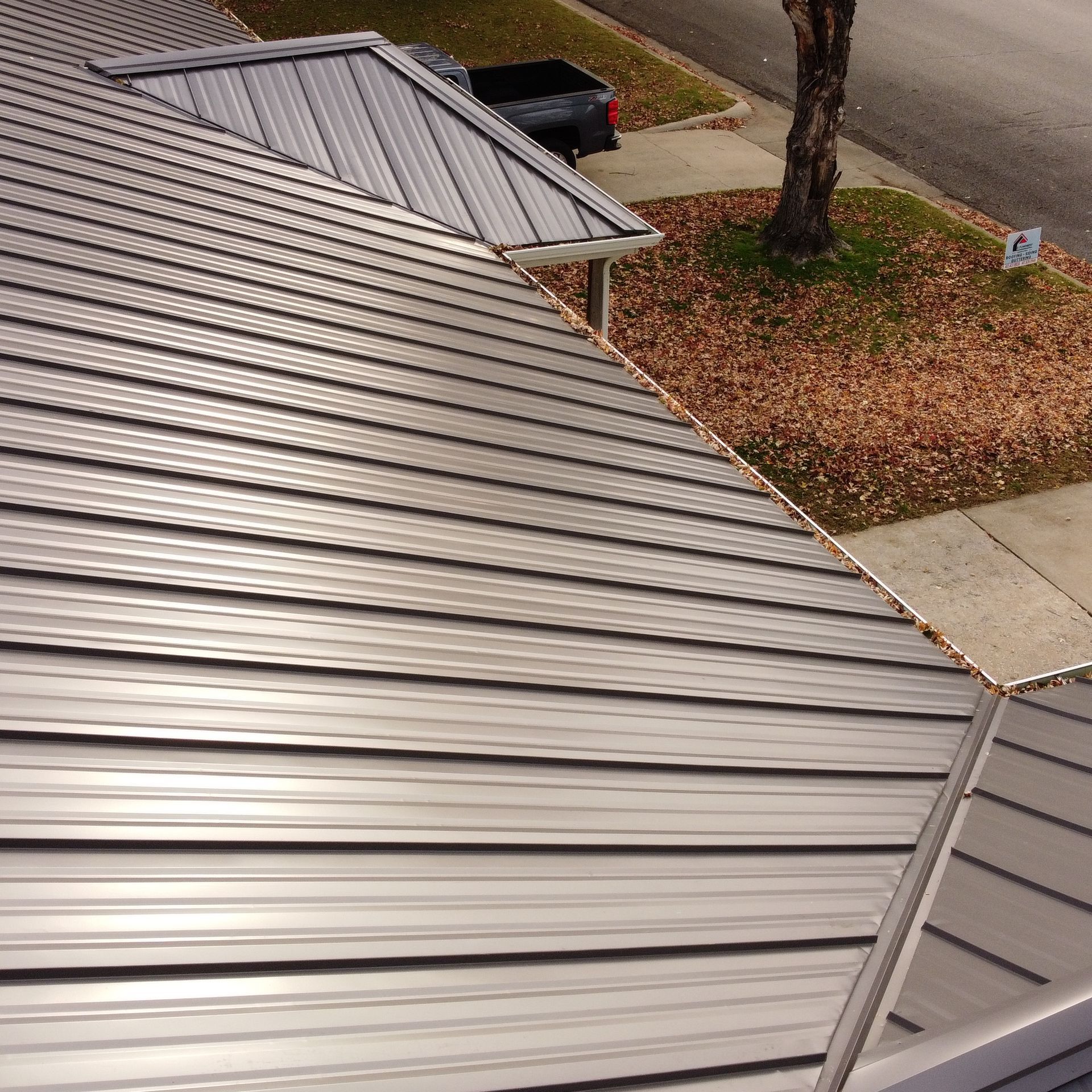 High-angle view of a grey metal roof with standing seams, transitioning toward a suburban yard and street.