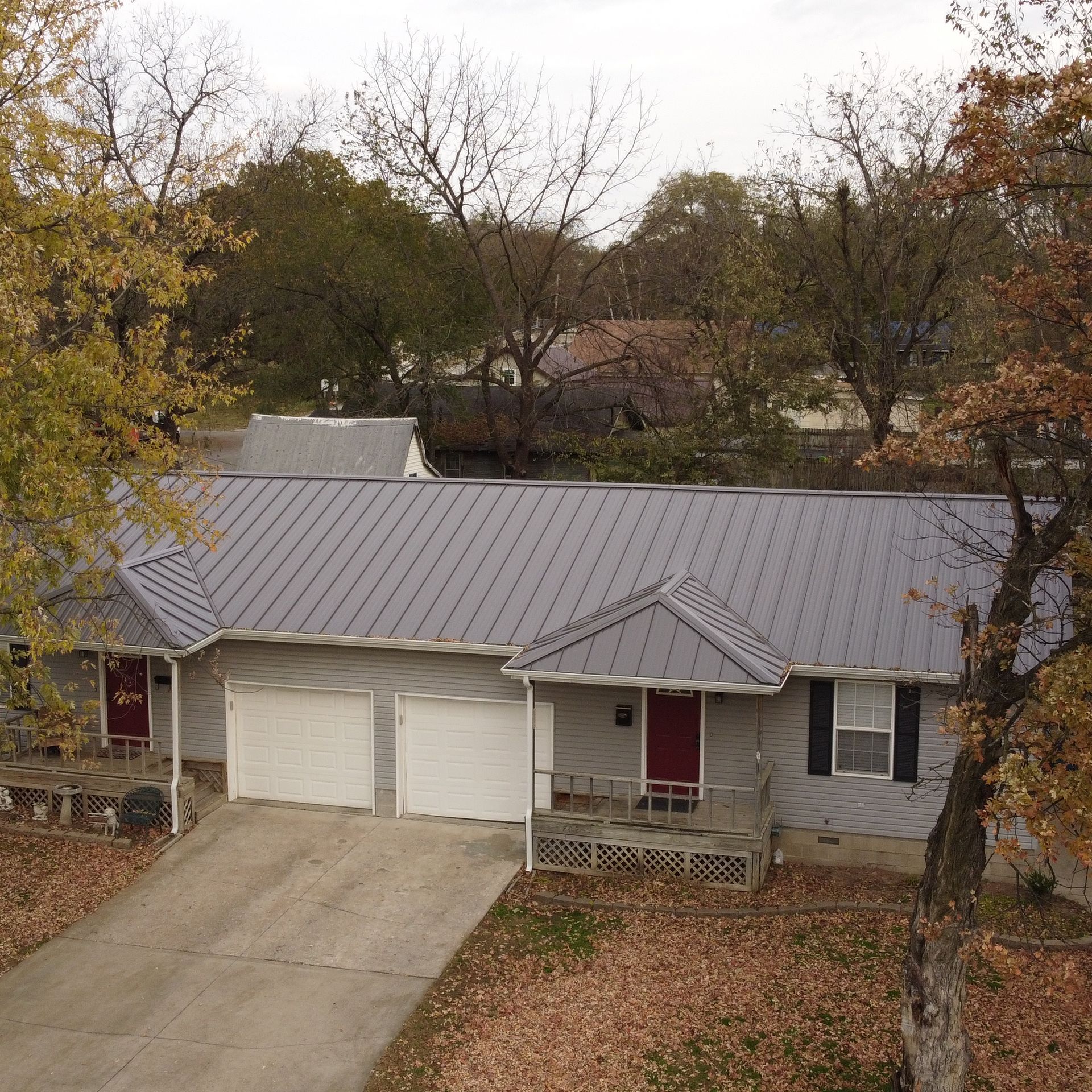 A single-story duplex with a gray metal roof, light siding, two white garage doors, and red front doors amid autumn trees.