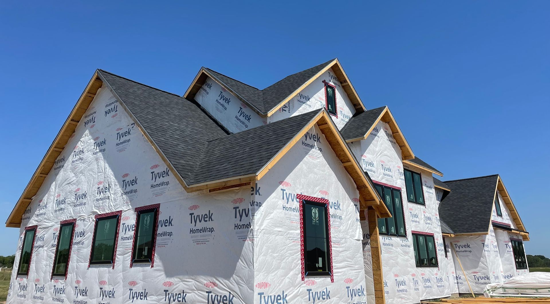 A new suburban house under construction, featuring dark shingles and white Tyvek house wrap against a clear blue sky.