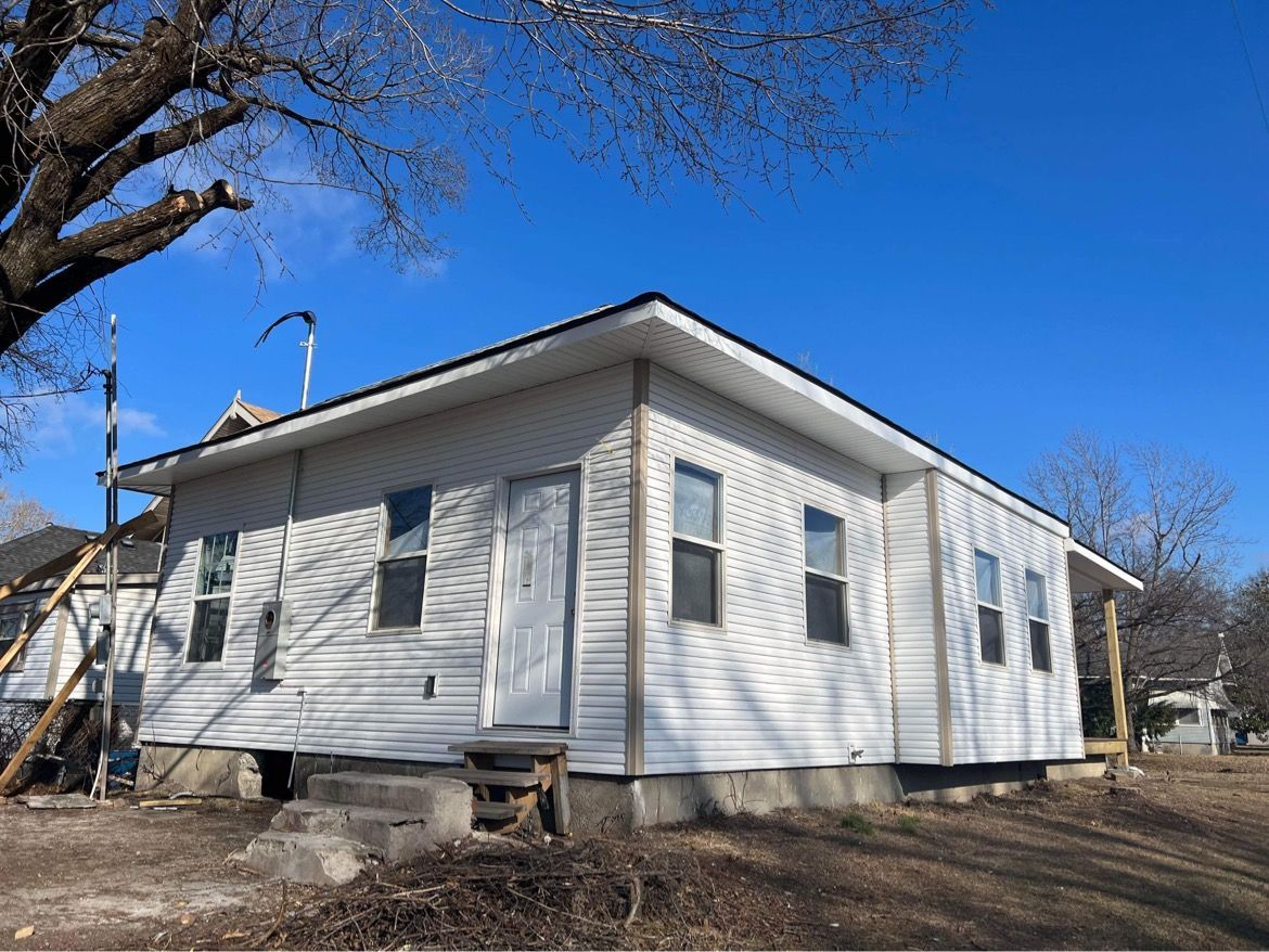 A white, single-story house with horizontal siding and a low-pitched roof, viewed on a sunny day with a clear blue sky.