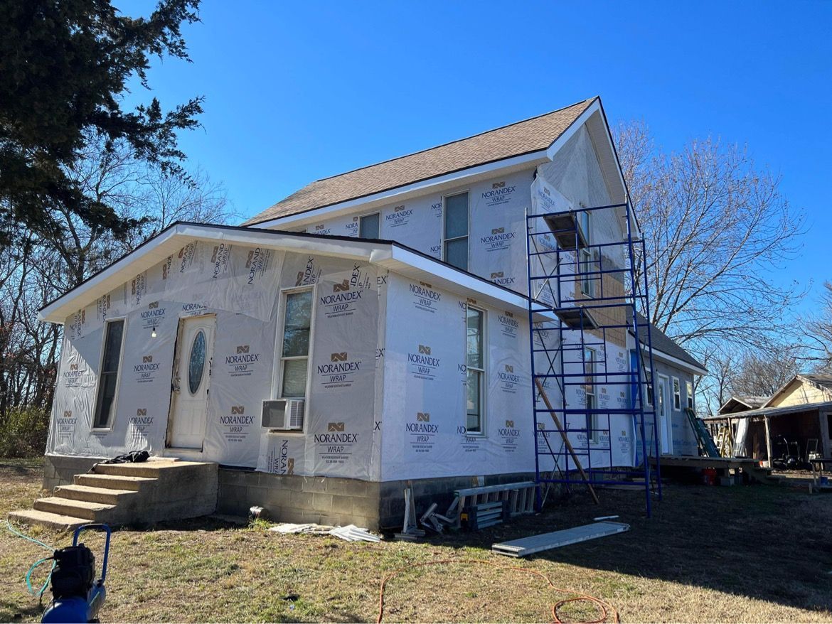 A house under construction wrapped in white house wrap, featuring blue scaffolding on the side under a clear blue sky.