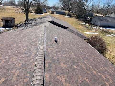 A high-angle view of a residential asphalt shingle roof with a central ridge cap, a vent pipe, and a distant backyard.