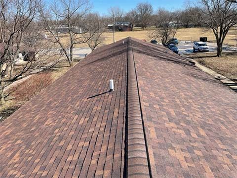 A view looking down the ridge of a brown asphalt shingle roof on a sunny day with trees and a parking lot in the distance.