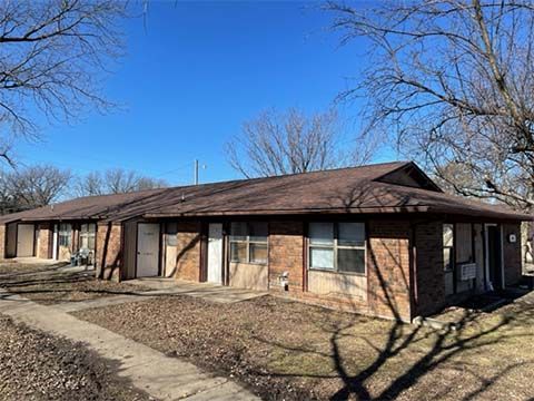 A long, single-story brick apartment building under a clear blue sky, surrounded by bare trees and dry, brown grass.