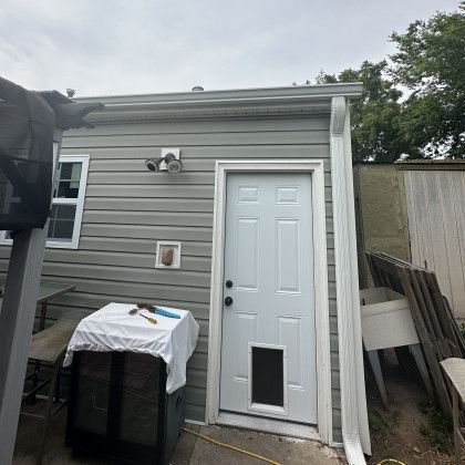 The exterior of a house with grey siding, a white door featuring a pet door, a window, and a white downspout.