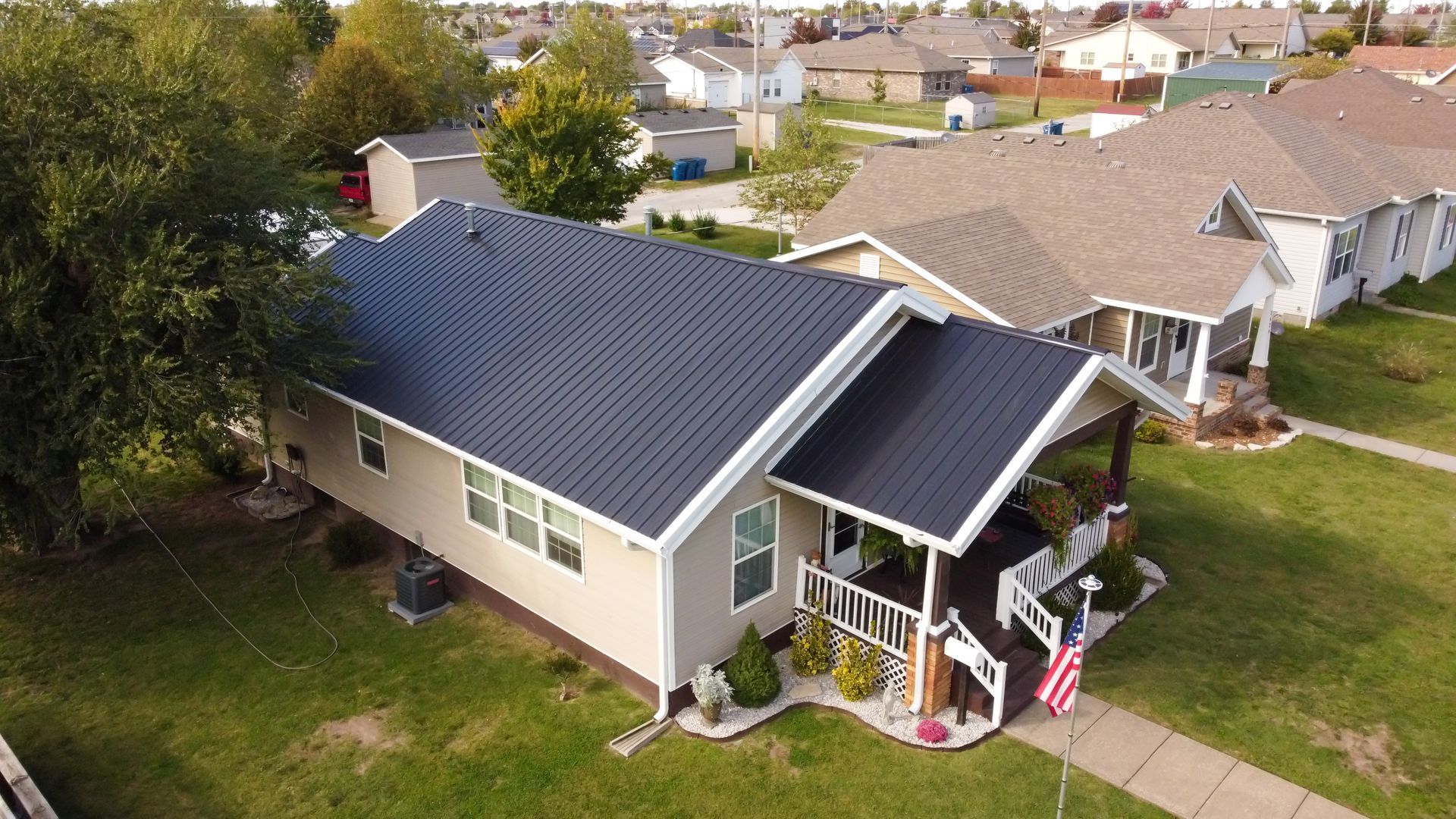 Aerial view of a single-story beige house with a dark metal roof, a front porch, and a small American flag in the yard.