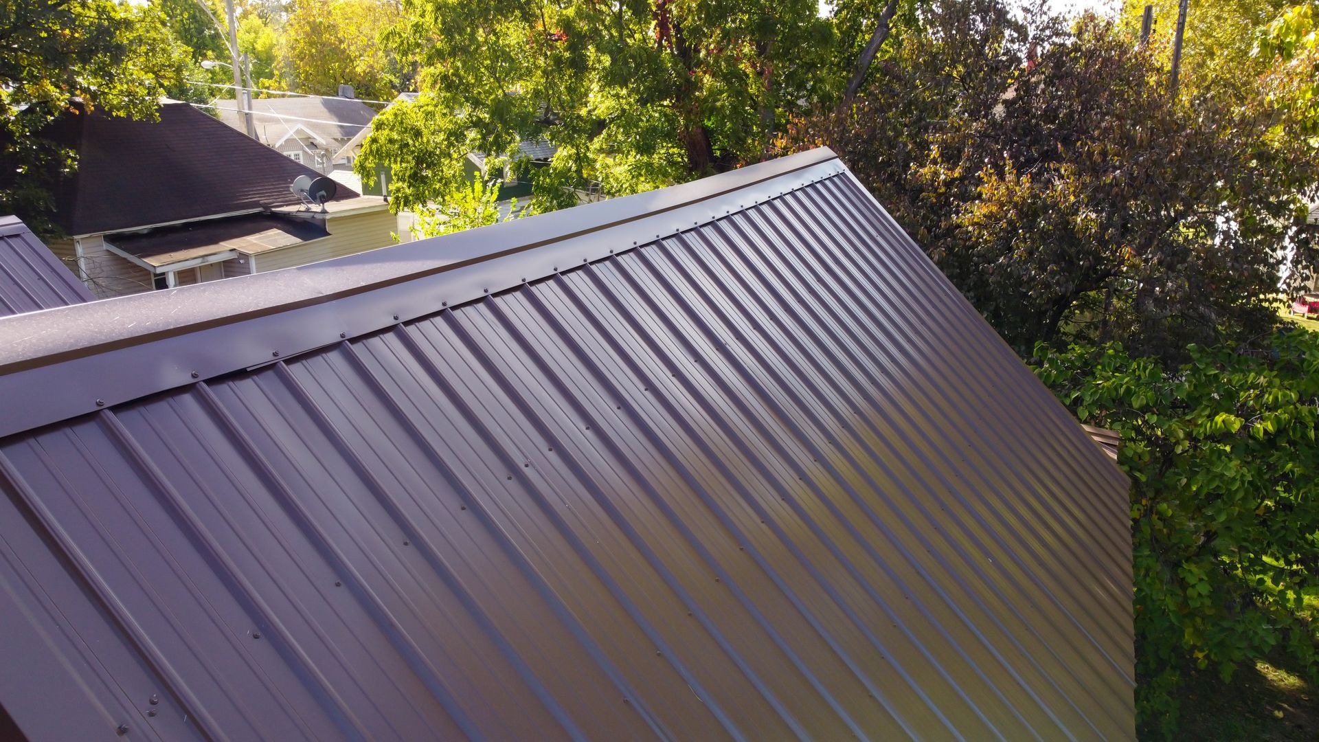 A high-angle view of a brown metal roof with vertical ribbed panels and a ridge cap, set against trees and a house.