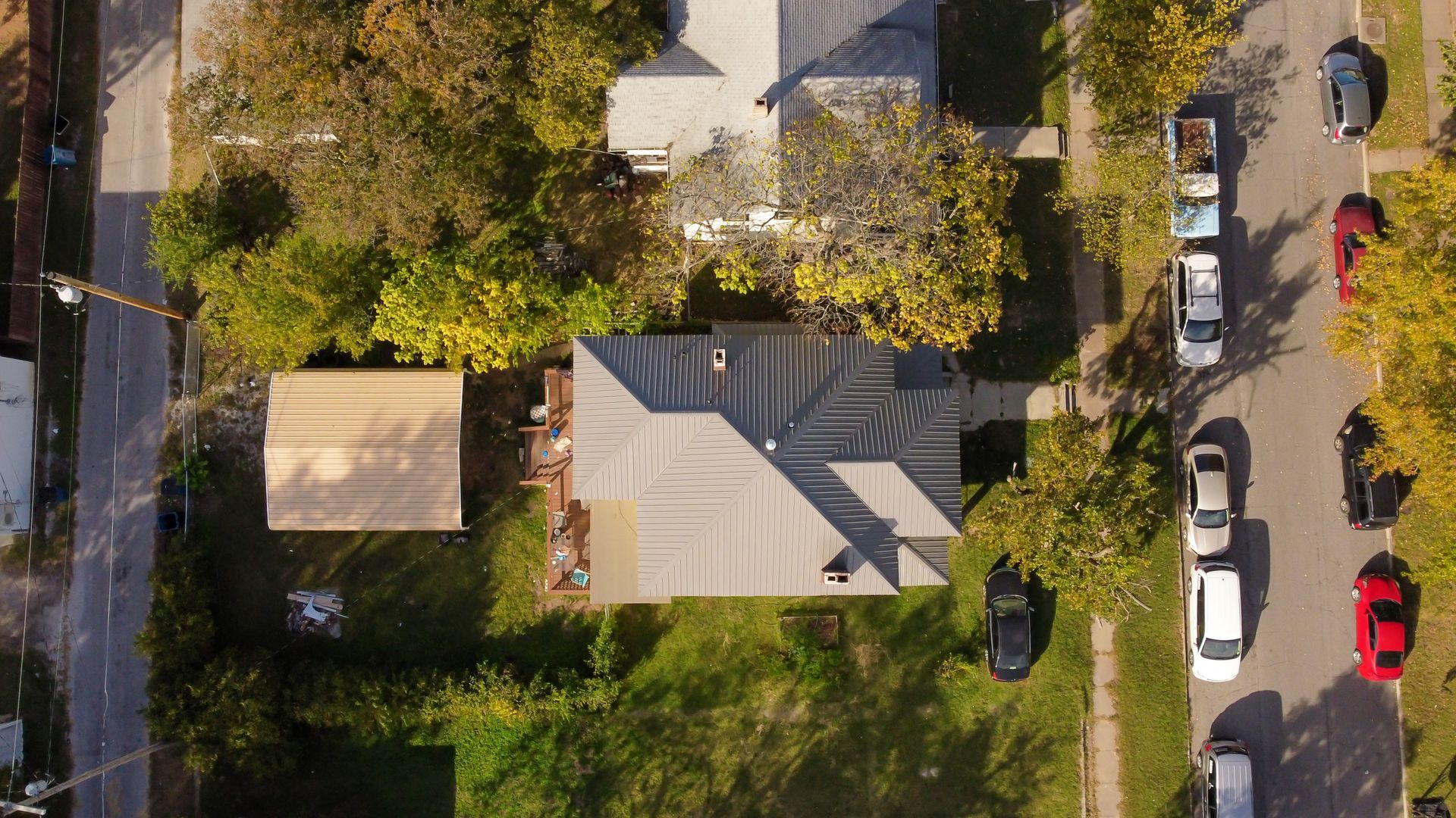 An aerial view of a suburban house, a detached carport, and parked cars on a street lined with green trees.