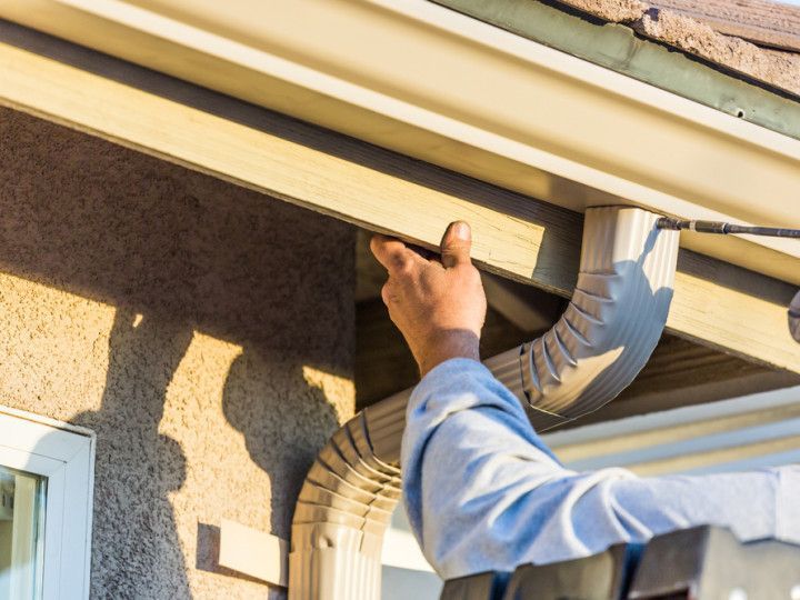 A worker installs a metal downspout onto the white gutter system of a stucco house.