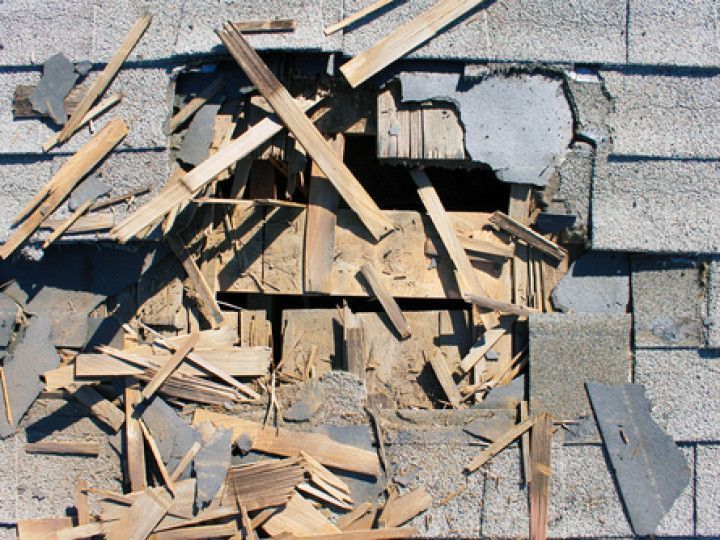A damaged residential roof with a hole exposing wooden rafters, surrounded by shattered shingles and splintered wood.