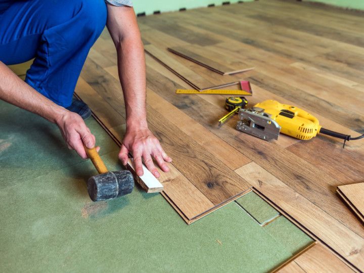 A person in blue pants using a mallet to tap a wood flooring plank into place on a green underlayment.