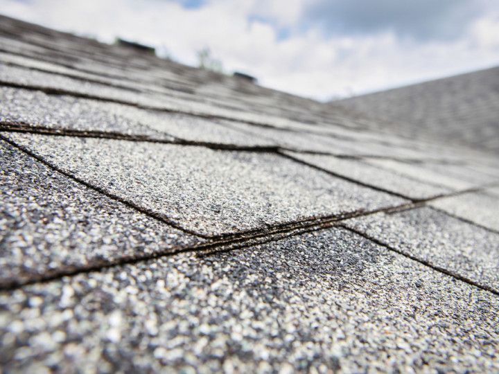 Close-up of gray asphalt roof shingles layered on a sloped roof under a cloudy sky.