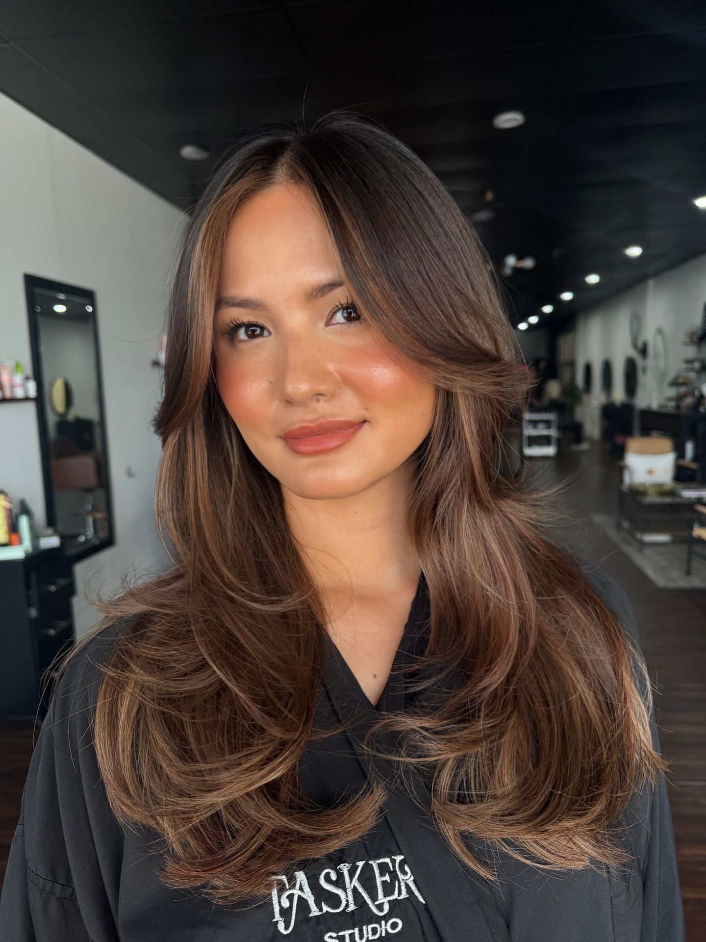 Woman with long layered brown hair and face-framing bangs smiles indoors, in a salon setting.