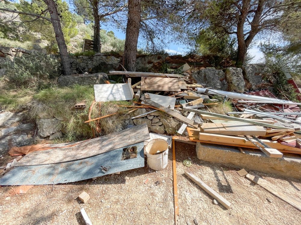 Piles of lumber and scrap metal scattered outdoors among rocks and pine trees