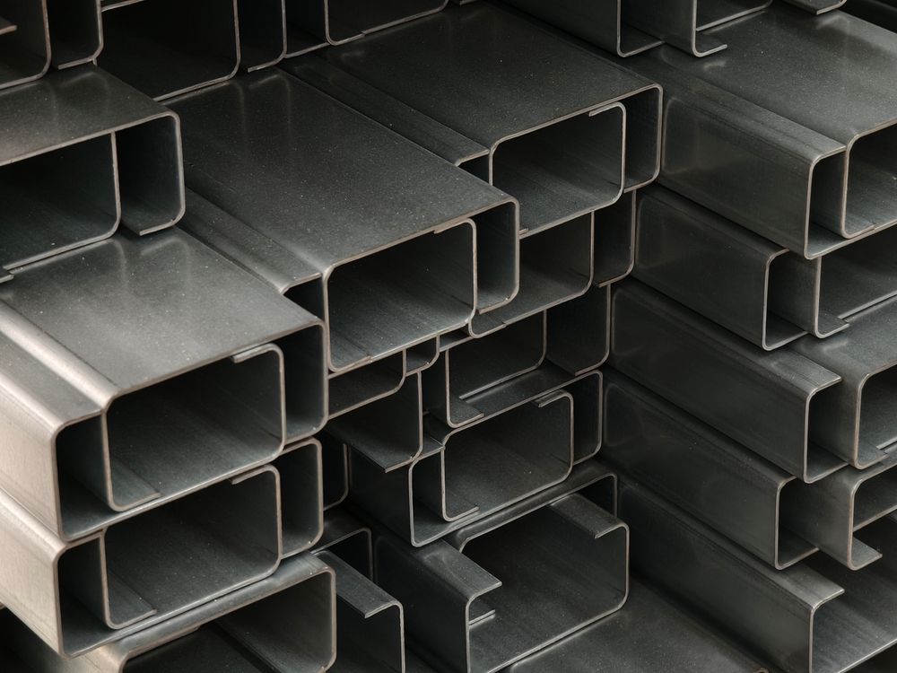 Stacks of dark gray metal rectangular tubes with hollow ends, piled in a warehouse-like setting