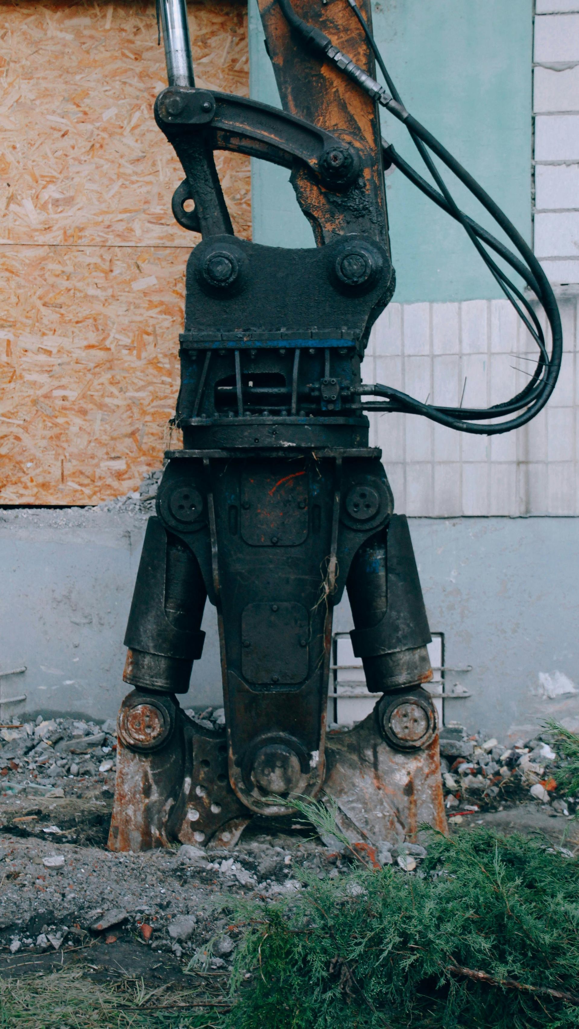 Old, rusted industrial machine standing outdoors beside a wall, with cables and overgrown weeds nearby