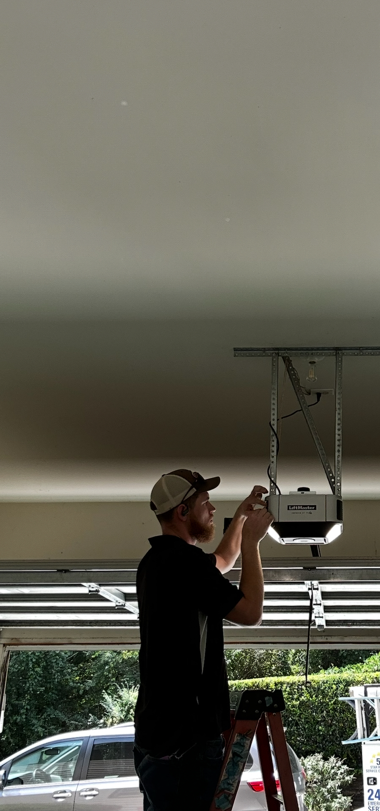 A person stands on a ladder, installing or repairing a garage door opener mounted to the ceiling.