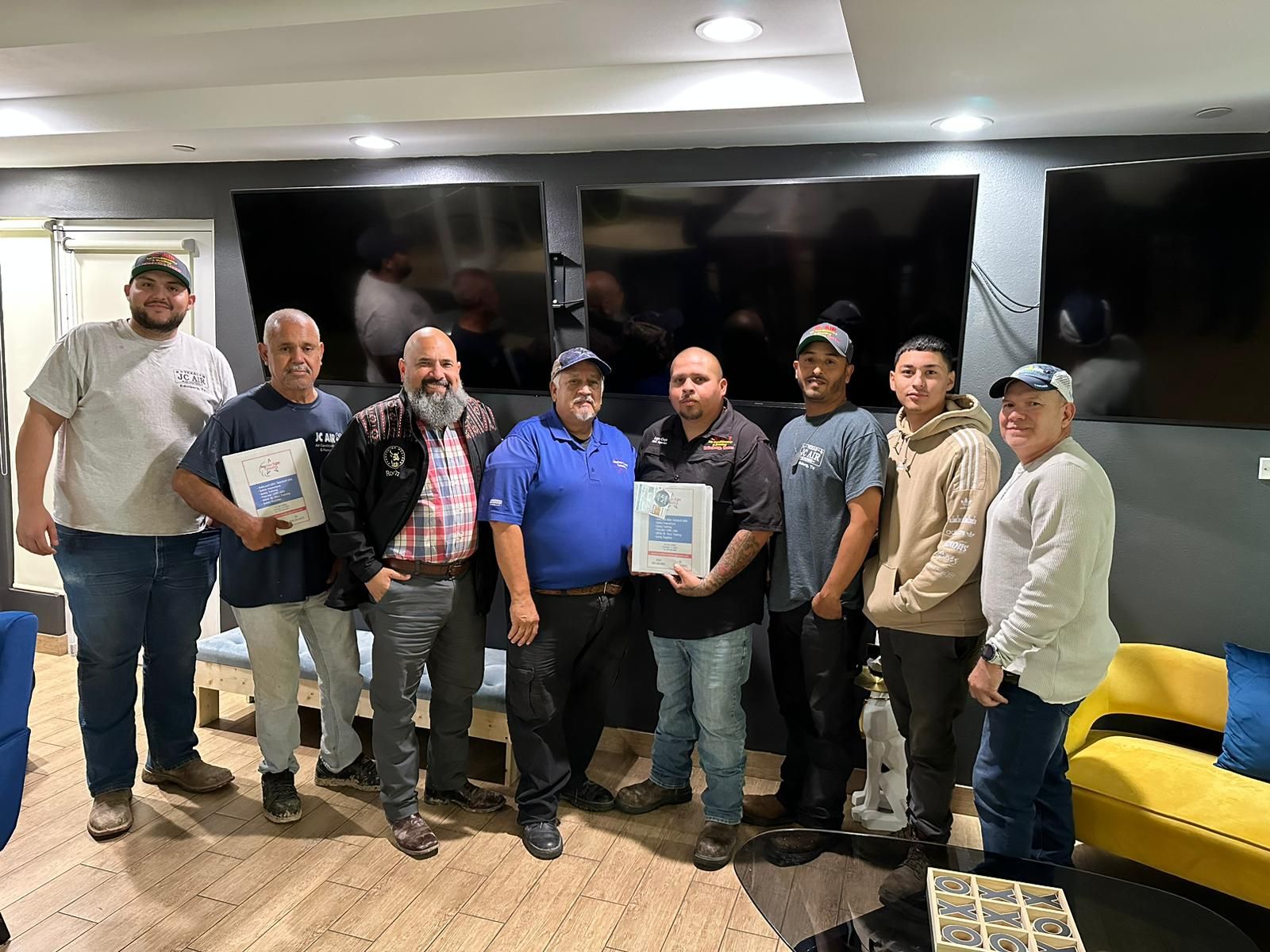 Group of men holding certificates, smiling in a room with large TVs and seating.