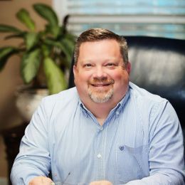 A man in a striped shirt is sitting at a table with a plant in the background.