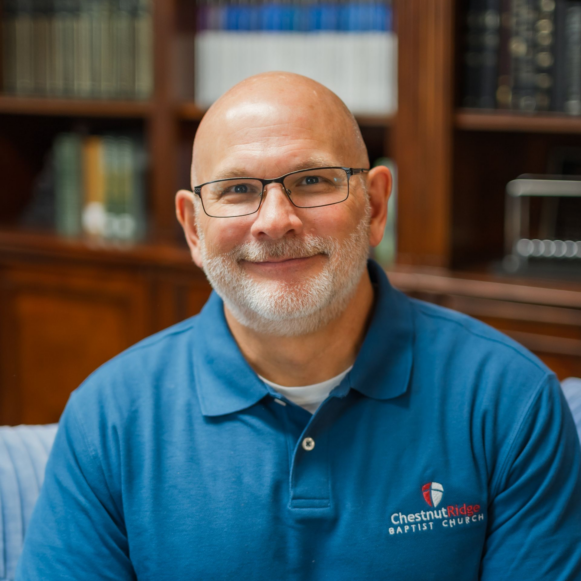 A bald man with a beard wearing glasses and a maroon shirt is sitting in front of a bookshelf.