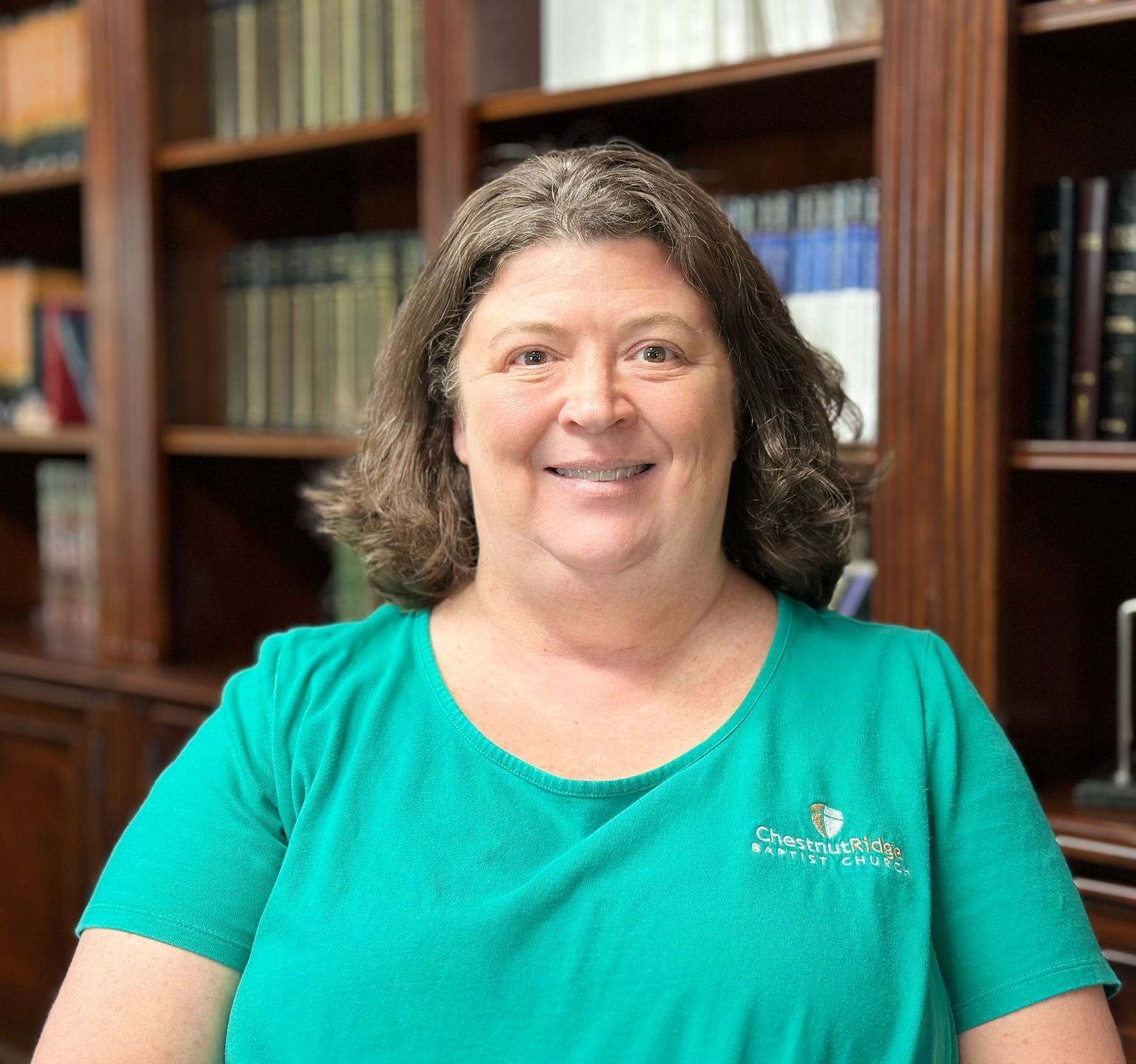 A woman in a green shirt is smiling in front of a bookshelf.