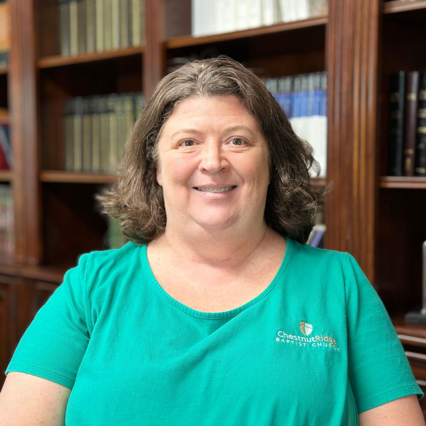 A woman in a green shirt is smiling in front of a bookshelf.