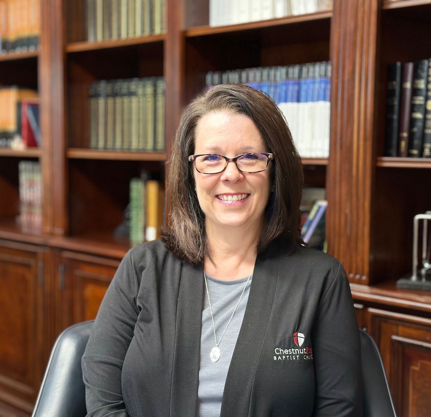 A woman wearing glasses and a black jacket is sitting in front of a bookshelf.