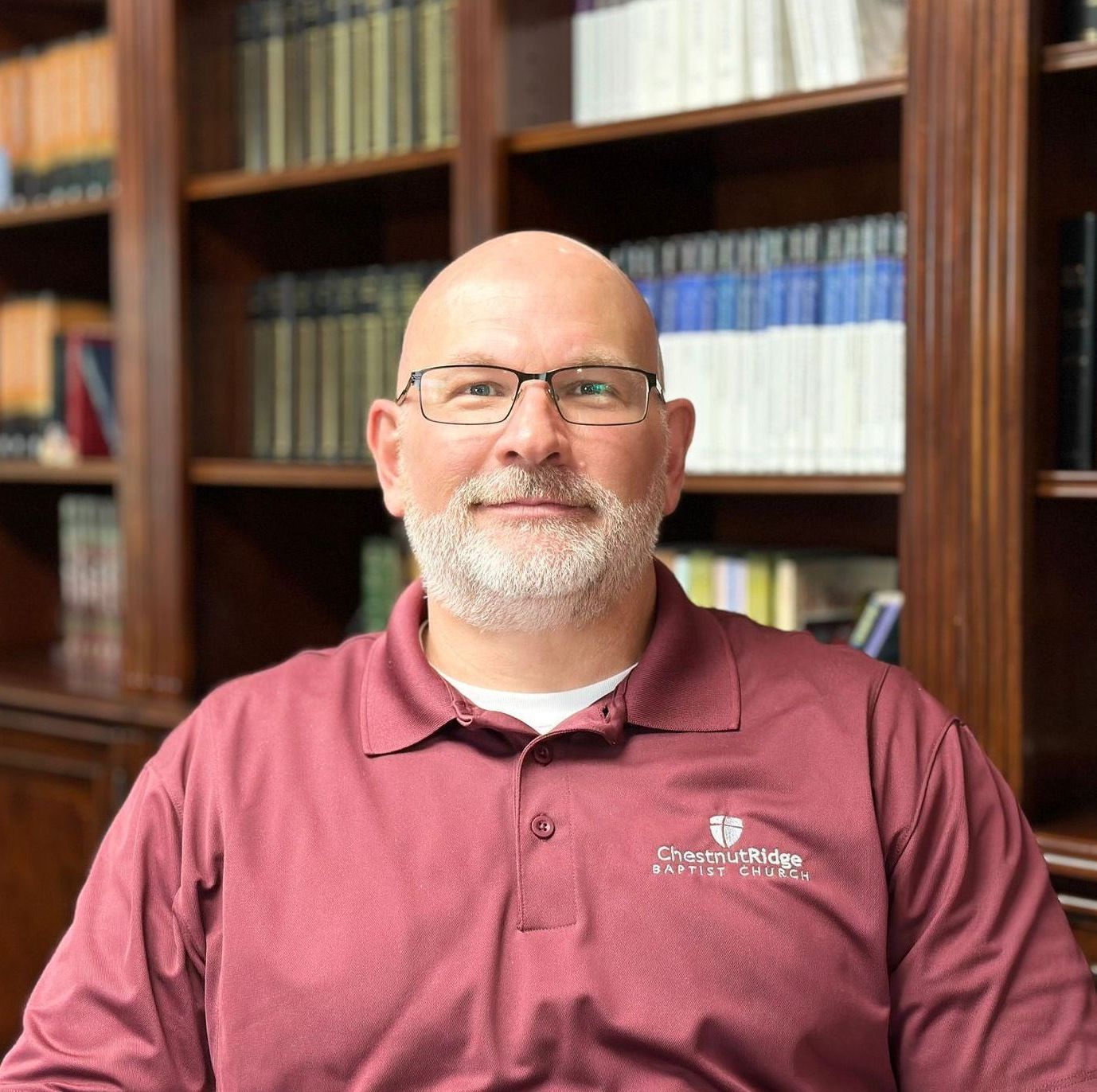 A bald man with a beard wearing glasses and a maroon shirt is sitting in front of a bookshelf.