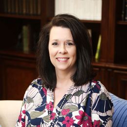 A woman in a floral shirt is smiling for the camera while sitting on a couch.