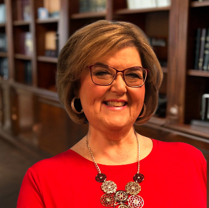 A woman wearing glasses and a red shirt is smiling in front of a bookshelf.