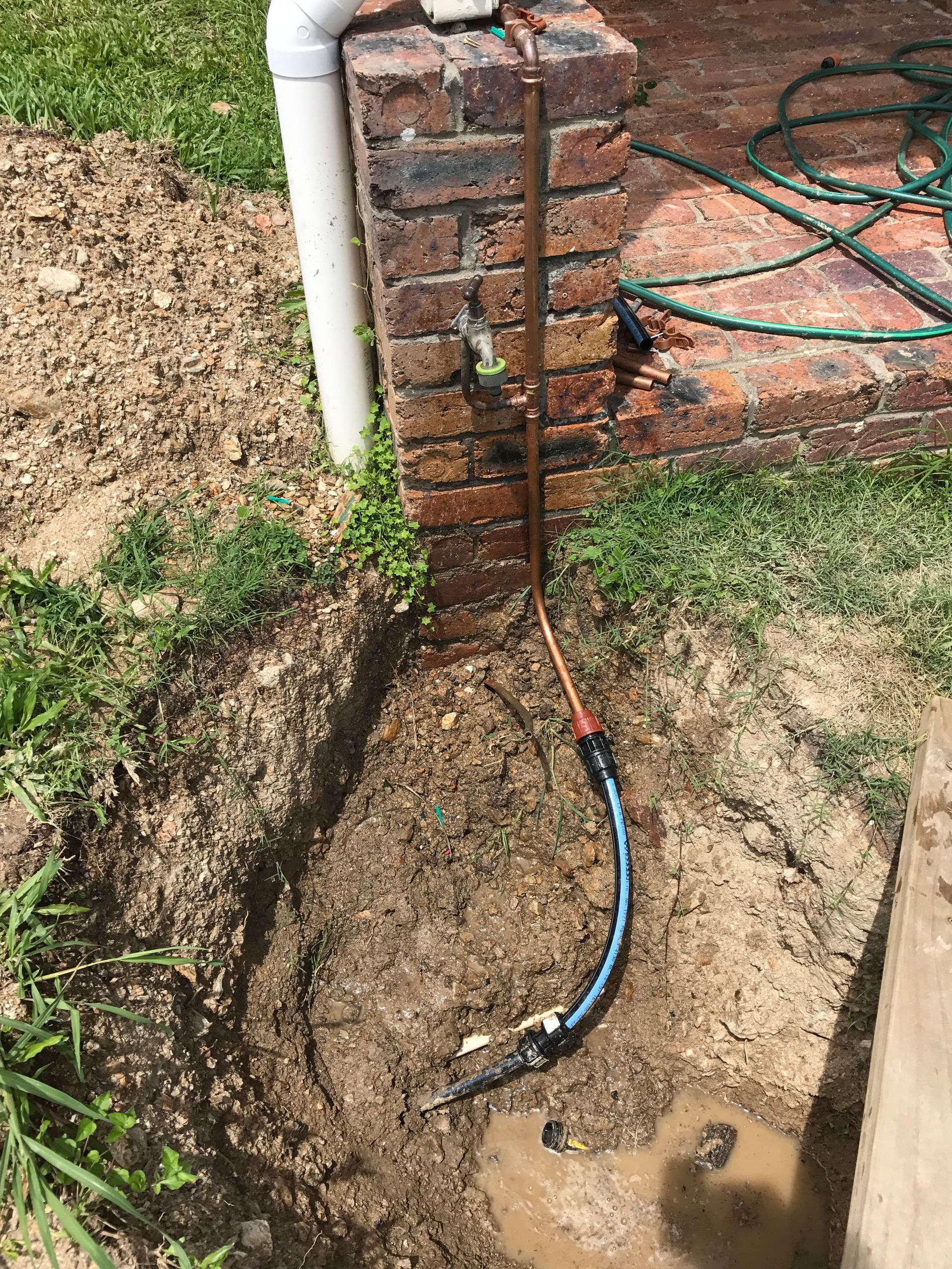 Copper pipe connected to black and blue hose in a hole in the ground next to a brick wall.